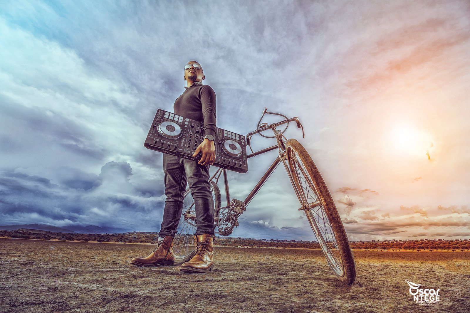 Moustey holding a DJ controller beside an old bicycle on an open plain under a dramatic sky